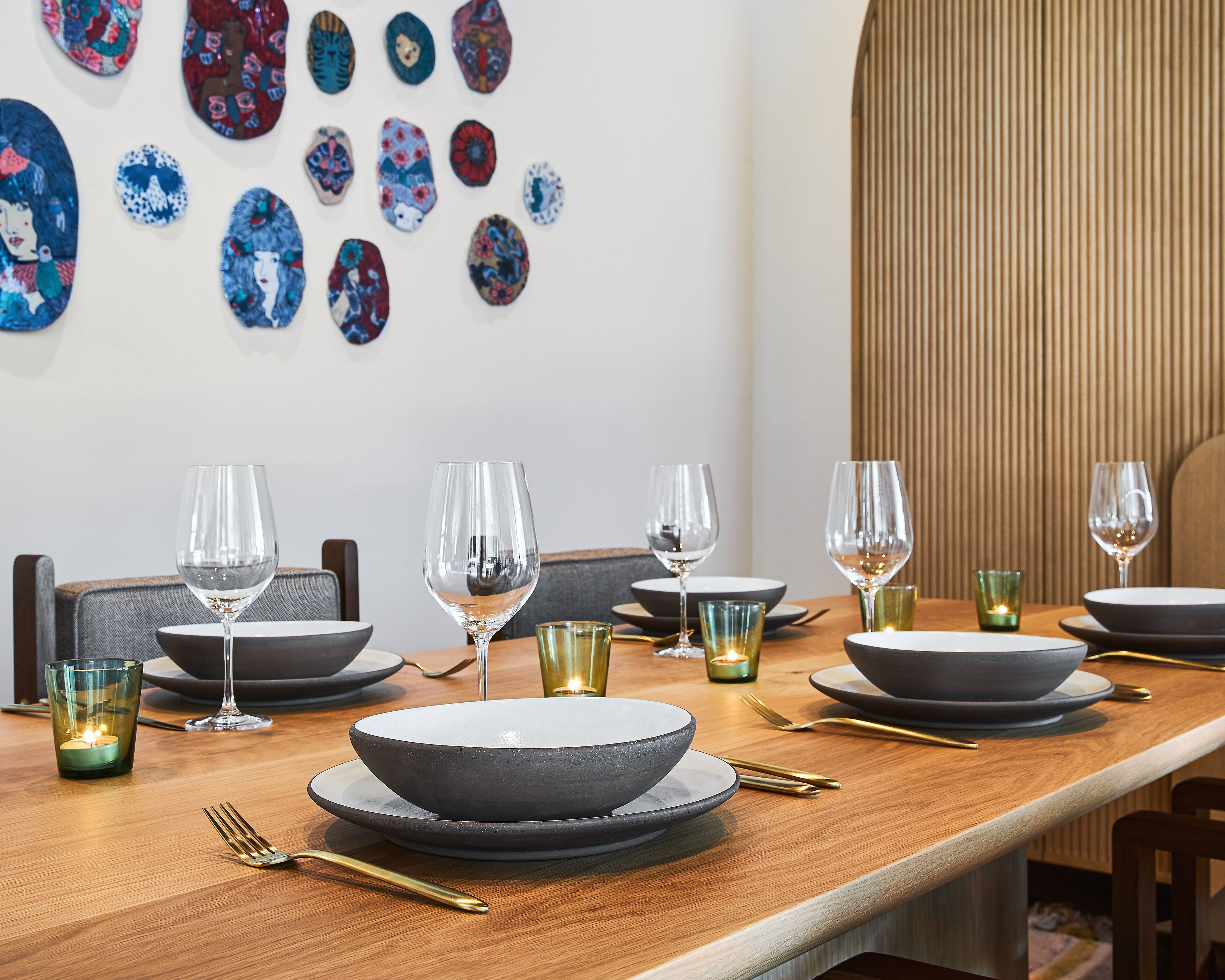 A dining table set with multiple bowls, glasses, and cutlery, with a candle and decorative plates on the wall behind.