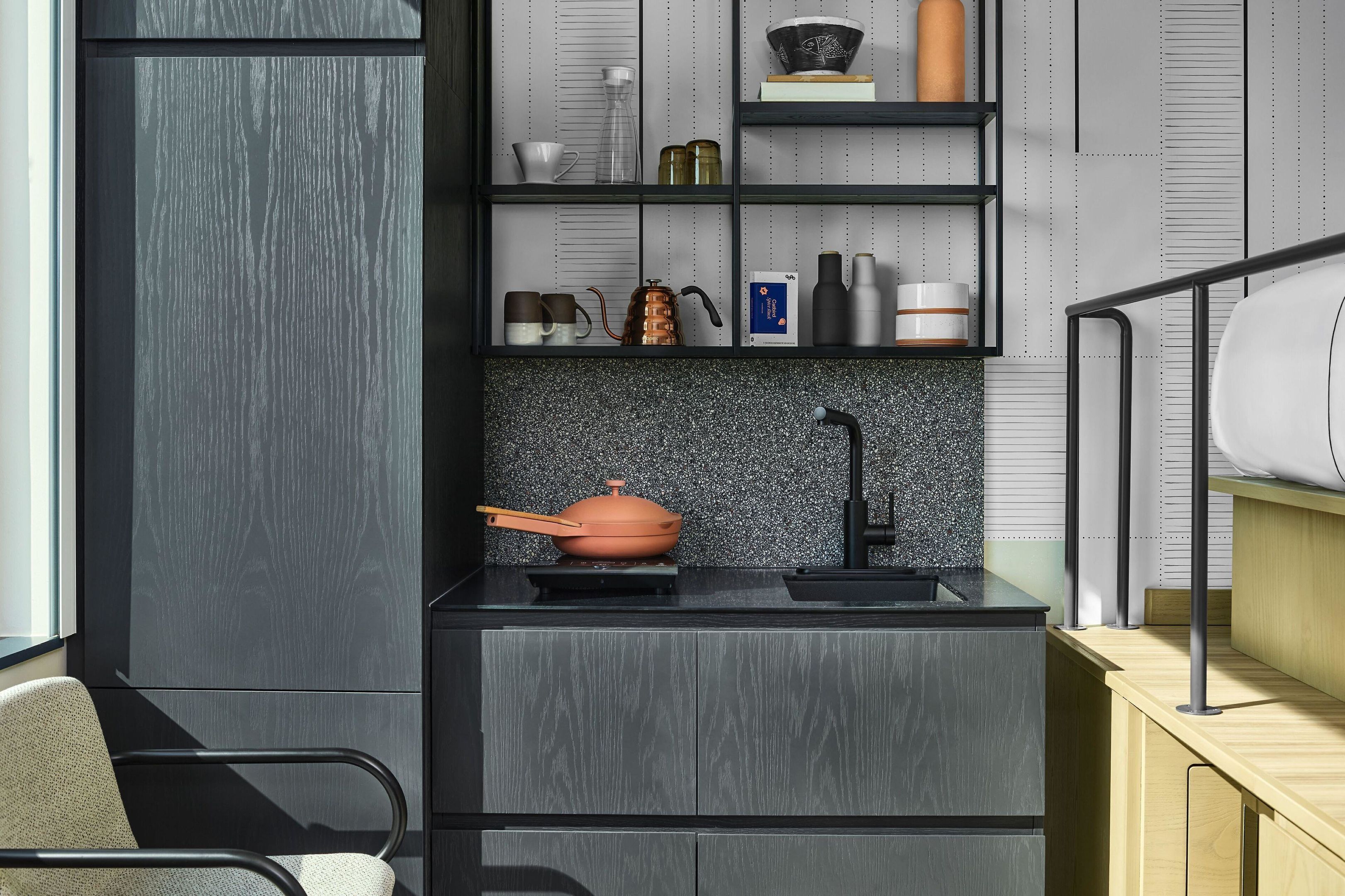 A modern kitchen with a black countertop, a stove, and a sink. There is a chair and a shelf with various kitchen items.