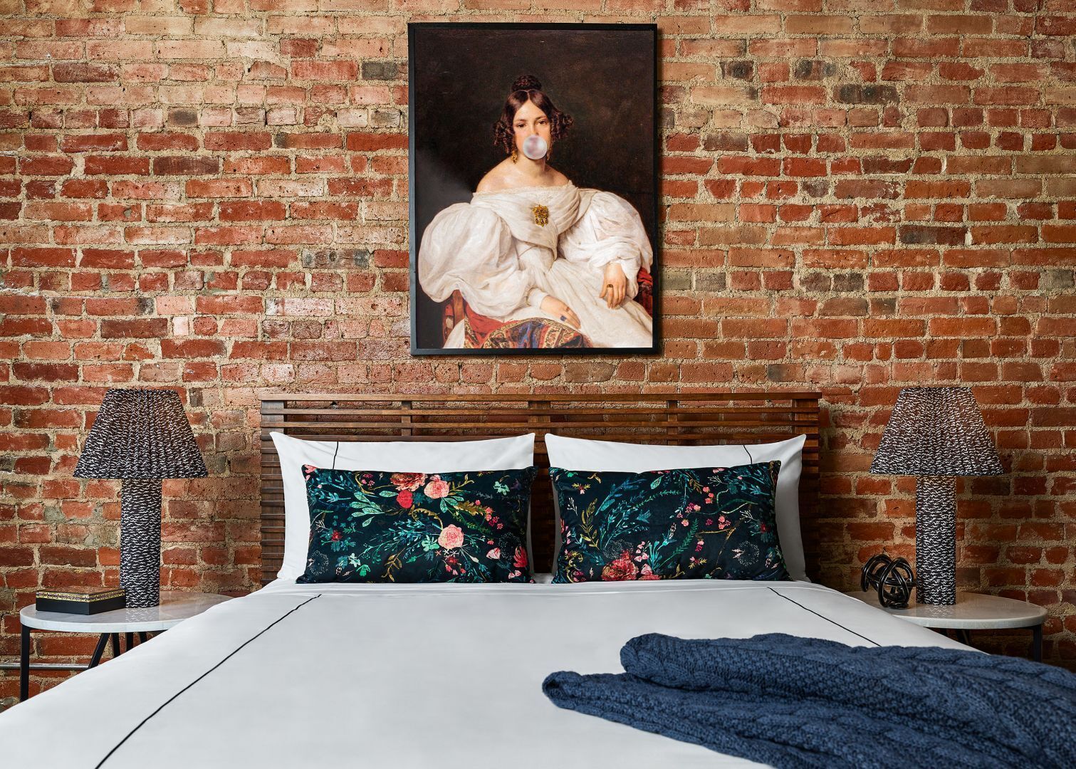 Un dormitorio con pared de ladrillo, una cama con almohadones florales y dos lámparas a cada lado.