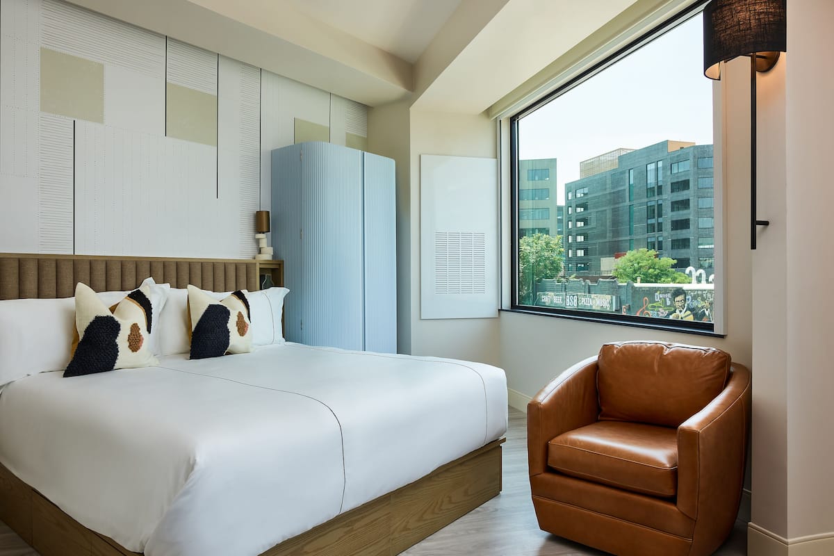 A modern hotel room with a large bed, a brown leather armchair, and a city view through a large window.