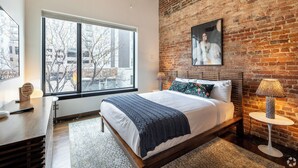 Un dormitorio moderno con un ventanal grande, una pared de ladrillo, una cama con una manta azul y una mesita de noche con una lámpara.