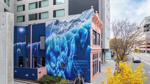 Un edificio con un mural de diseño abstracto en azul, una estatua de elefante y un árbol con hojas amarillas.