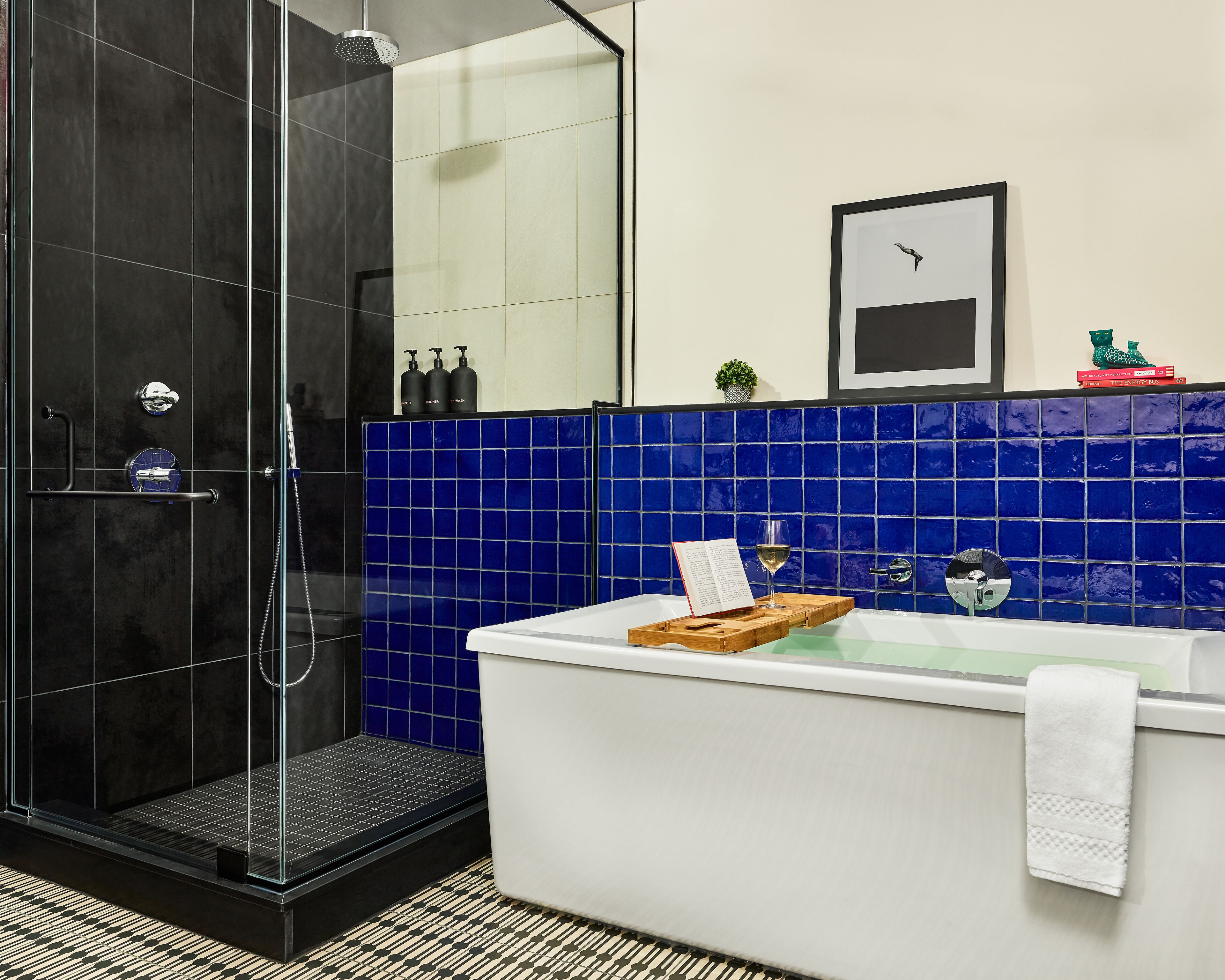 A modern bathroom with a glass-enclosed shower, a white bathtub with a wooden tray, and a blue-tiled wall.