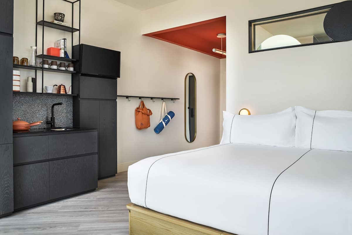 A modern hotel room with a bed, a kitchenette, and a mirror.