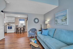 A living room with a sofa, a dining table with chairs, a kitchen area with a stove and microwave, and a wall-mounted mirror. - Odysea: Hampton Beach Getaway – Seafood, Music & Waves Await! (Hampton)