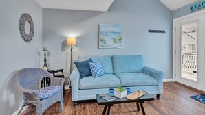 Una sala de estar con un sofá azul claro, un sillón de mimbre, una lámpara de pie y un cuadro con una playa.