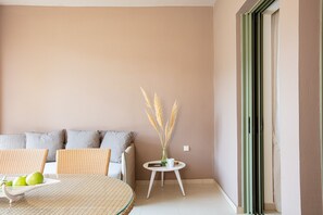 A room with a sofa, a round table with a basket of green apples, and a small white table with a vase of dried plants.