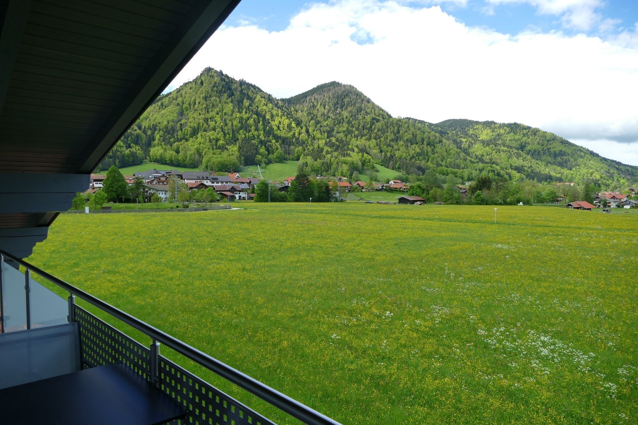 Vista desde un balcón sobre un campo verde con casas y montañas al fondo.
