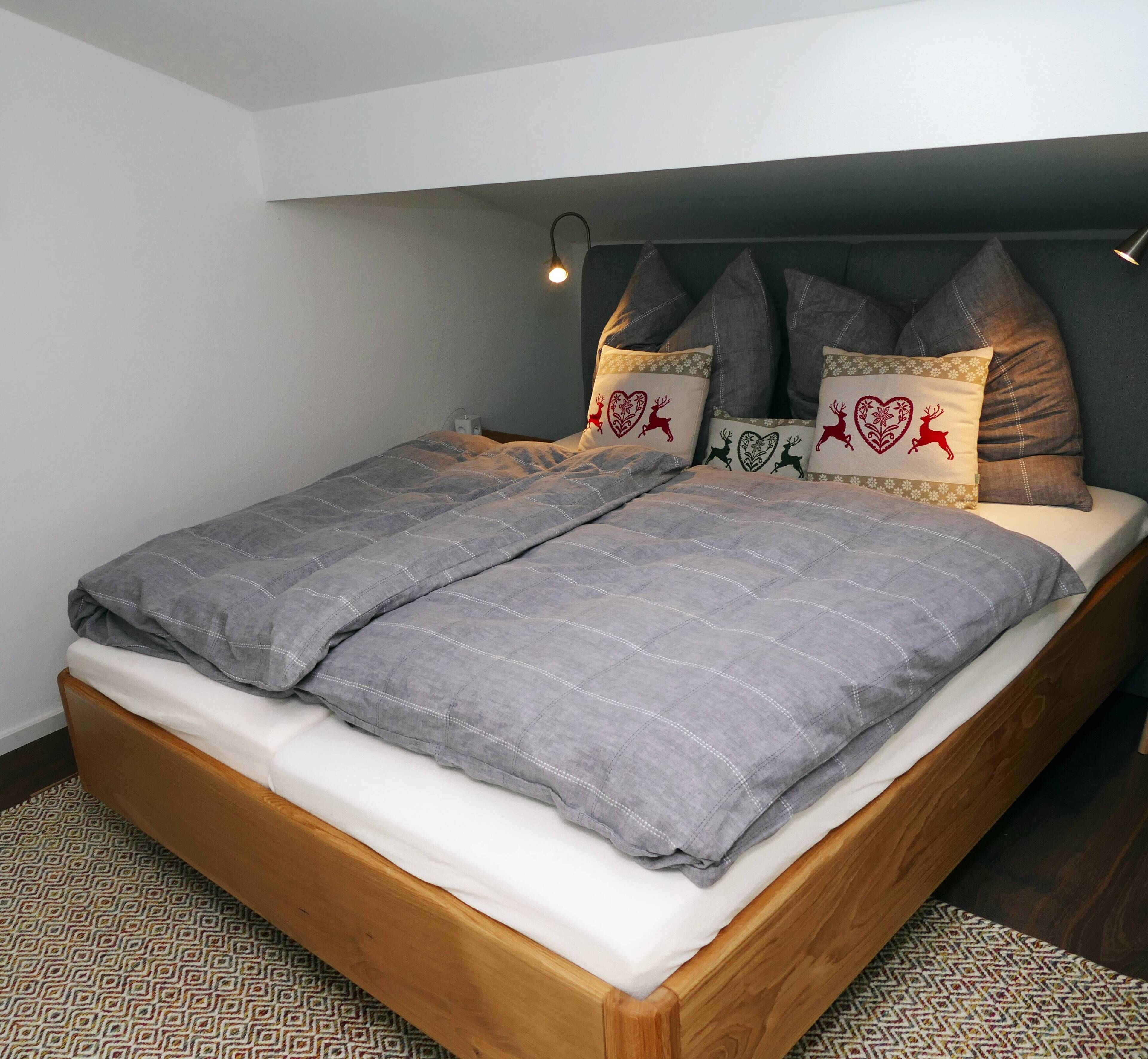 Ein ordentlich bezogenes Bett mit grauer Decke, einem Holzkopfende und dekorativen Kissen mit Herz- und Rentier-Motiven.