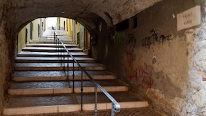 A staircase with metal railings, leading upwards through a tunnel-like structure with a sign reading 'VICOLO MANINI'.