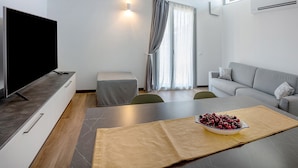 A modern living room with a flat-screen TV, a sofa, a coffee table with a bowl of fruit, and a dining area with a table and chairs.
