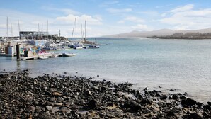 A rocky shoreline with boats docked at a marina, a building with a sloped roof, and mountains in the background. - Apartment "Casa Raquel 1" with Sea View, Pool, Wi-Fi & Terrace (Costa Antigua)