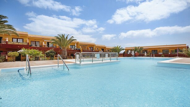 A swimming pool with a clear blue sky, palm trees, and yellow buildings with balconies. - Apartment "Casa Raquel 1" with Sea View, Pool, Wi-Fi & Terrace (Costa Antigua)