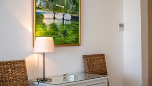 Ein Zimmer mit einem Gemälde von Booten an der Wand, einer Lampe, Korbstühlen und einem Glastisch.