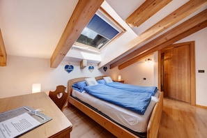 Een slaapkamer met dakraam, houten balken, een bed met blauw beddengoed, een nachtkastje en een houten deur.