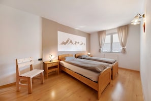 A bedroom with two wooden beds, a desk, a chair, a wall-mounted lamp, and a window with curtains.