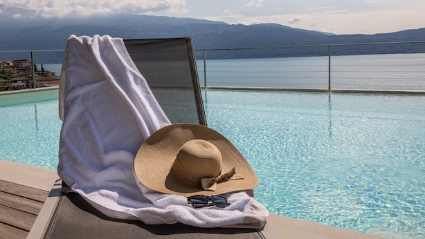 A poolside lounge chair with a towel, a straw hat, and sunglasses, overlooking a clear pool and distant mountains. - Exclusive Villa Agave with Lake & Mountain View, Pool, Whirlpool, and Sauna (Toscolano-Maderno)
