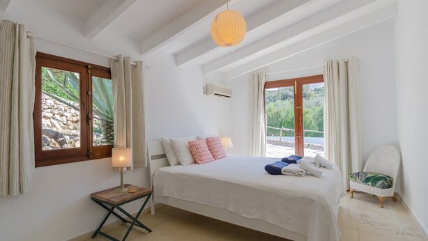 A bedroom with a bed, a bedside table with a lamp, a chair, and a view of greenery outside. - Vacation Villa "Villa Diani" with Pool, Wi-Fi, Garden & Terrace (Binibequer)
