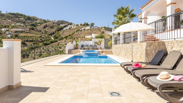 A pool area with lounge chairs, a stone wall, and a view of a hillside with houses. - Villa "Natalia" with Sea & Mountain View, Private Pool, Air Conditioning & Terrace (Nerja)