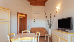 A dining area with a table and chairs, a sofa, a television on a cabinet, and a door leading to another room.