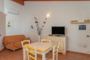 A room with a sofa, a dining table with chairs, a TV on a stand, and a wall-mounted shelf with decorative items.