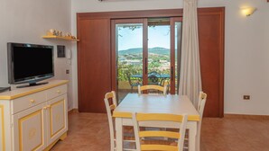 A room with a TV on a cabinet, a dining table with chairs, a sliding glass door to a balcony with a view, and a light fixture on the wall.