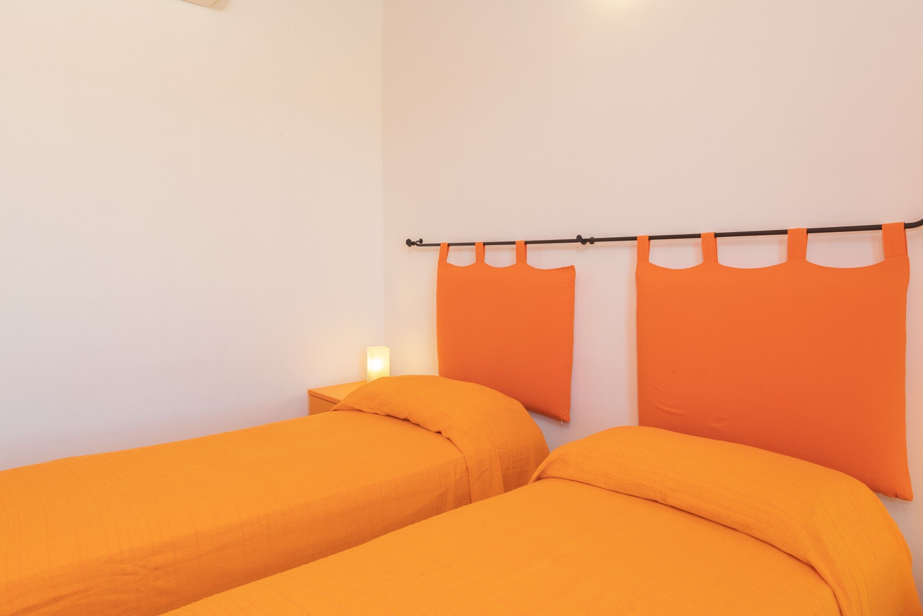 A room with two beds, each with orange bedding, a nightstand, and a wall-mounted curtain rod with orange curtains.