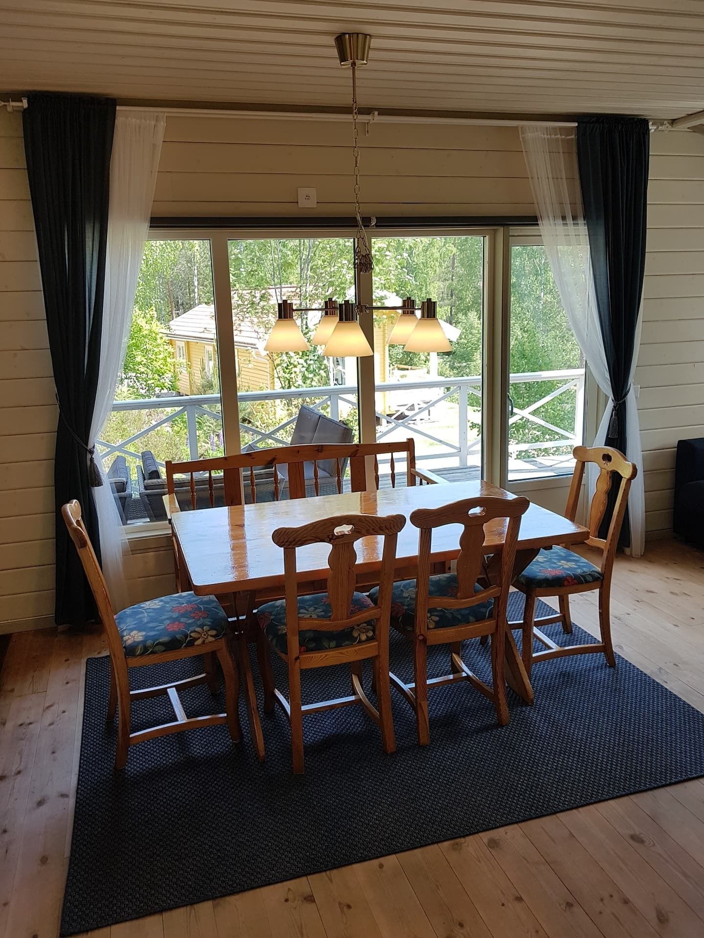 A dining area with a wooden table and chairs, a black rug, and a view of the outdoors through large windows.