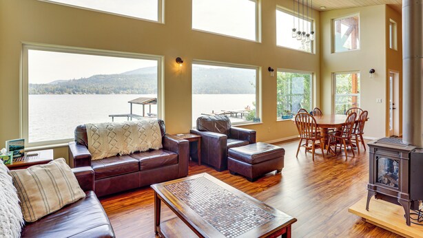 A spacious living room with a large window overlooking a lake, a cozy seating area, and a dining table with chairs. - Lake Whatcom House w/ Private Hot Tub + Dock (Bellingham)