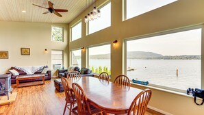 A spacious dining area with a large wooden table and chairs, a ceiling fan, and large windows overlooking a lake. - Lake Whatcom House w/ Private Hot Tub + Dock (Bellingham)
