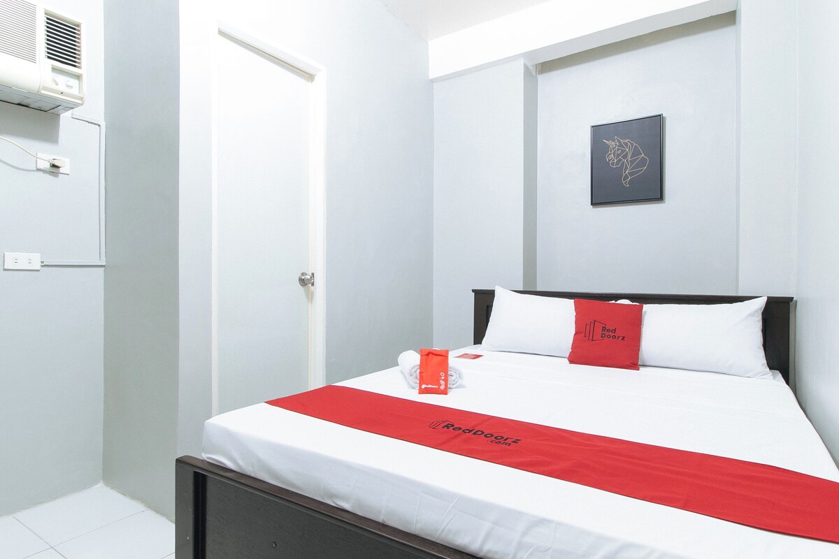 A hotel room with a bed, a red throw, a pillow with a logo, a framed picture on the wall, and an air conditioning unit.