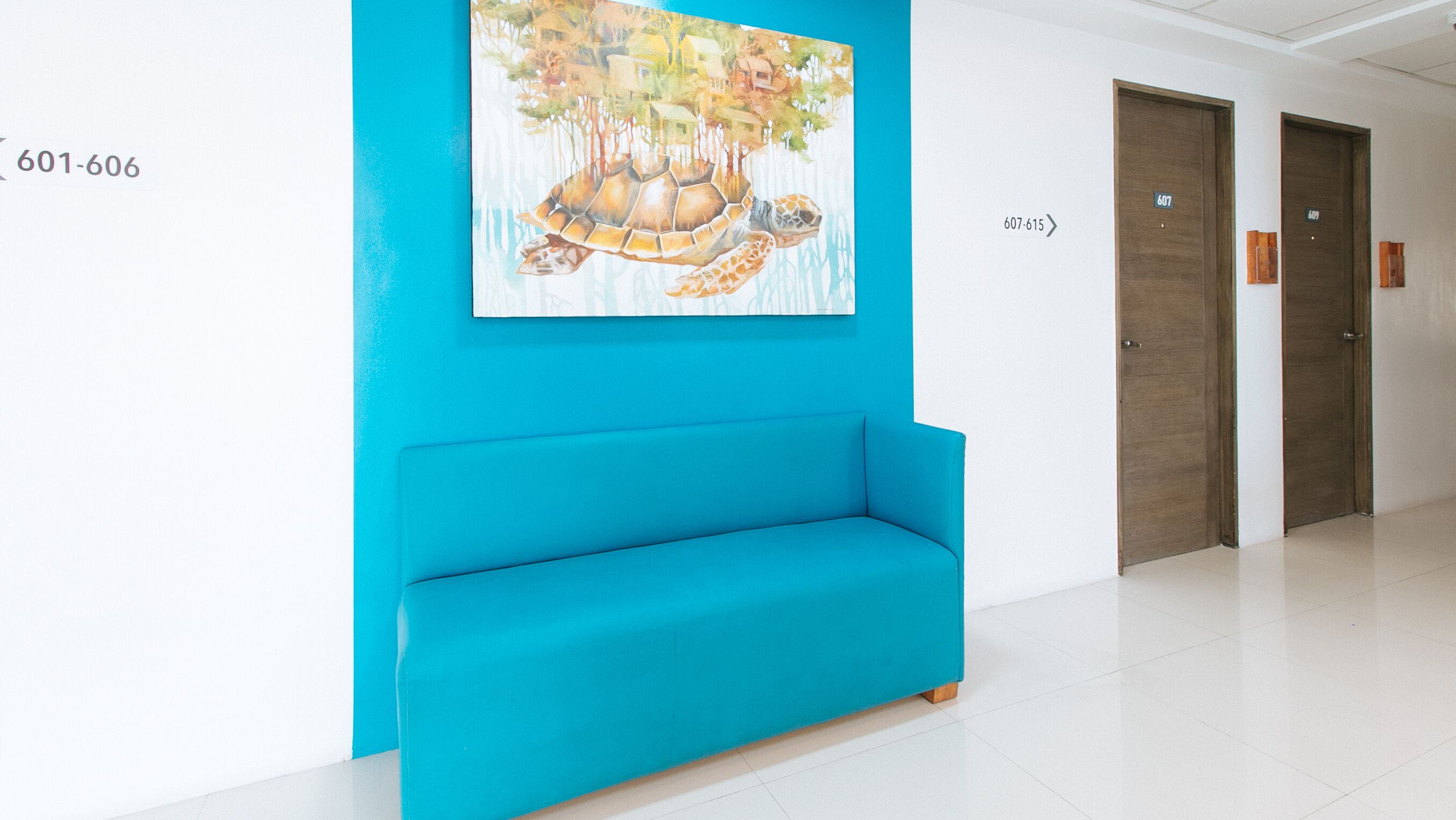 A bright blue bench in a hallway with a mural of a turtle on the wall and doors numbered 607-615.