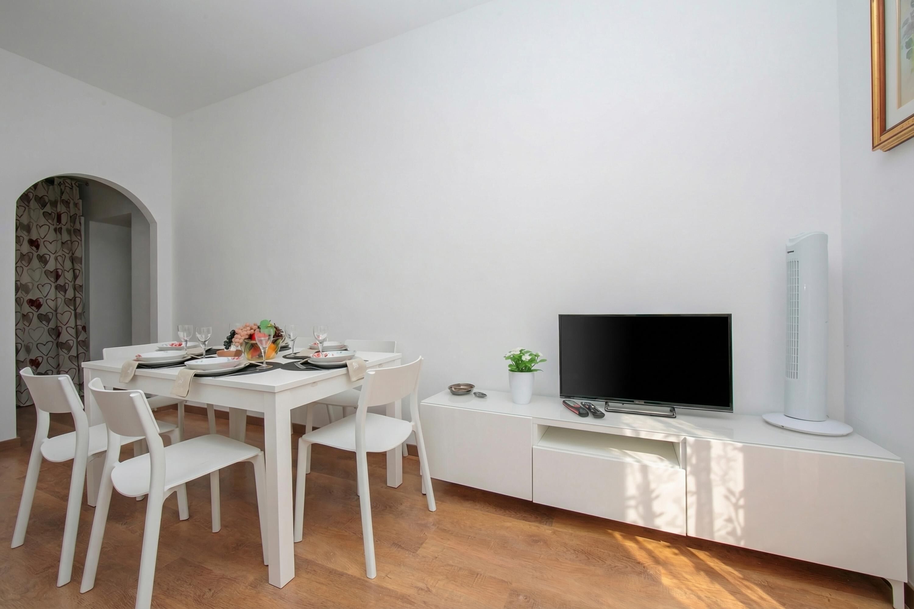 Ett modernt matområde med ett vitt bord för fyra personer, en platt-TV och ett vitt skåp med en växt.