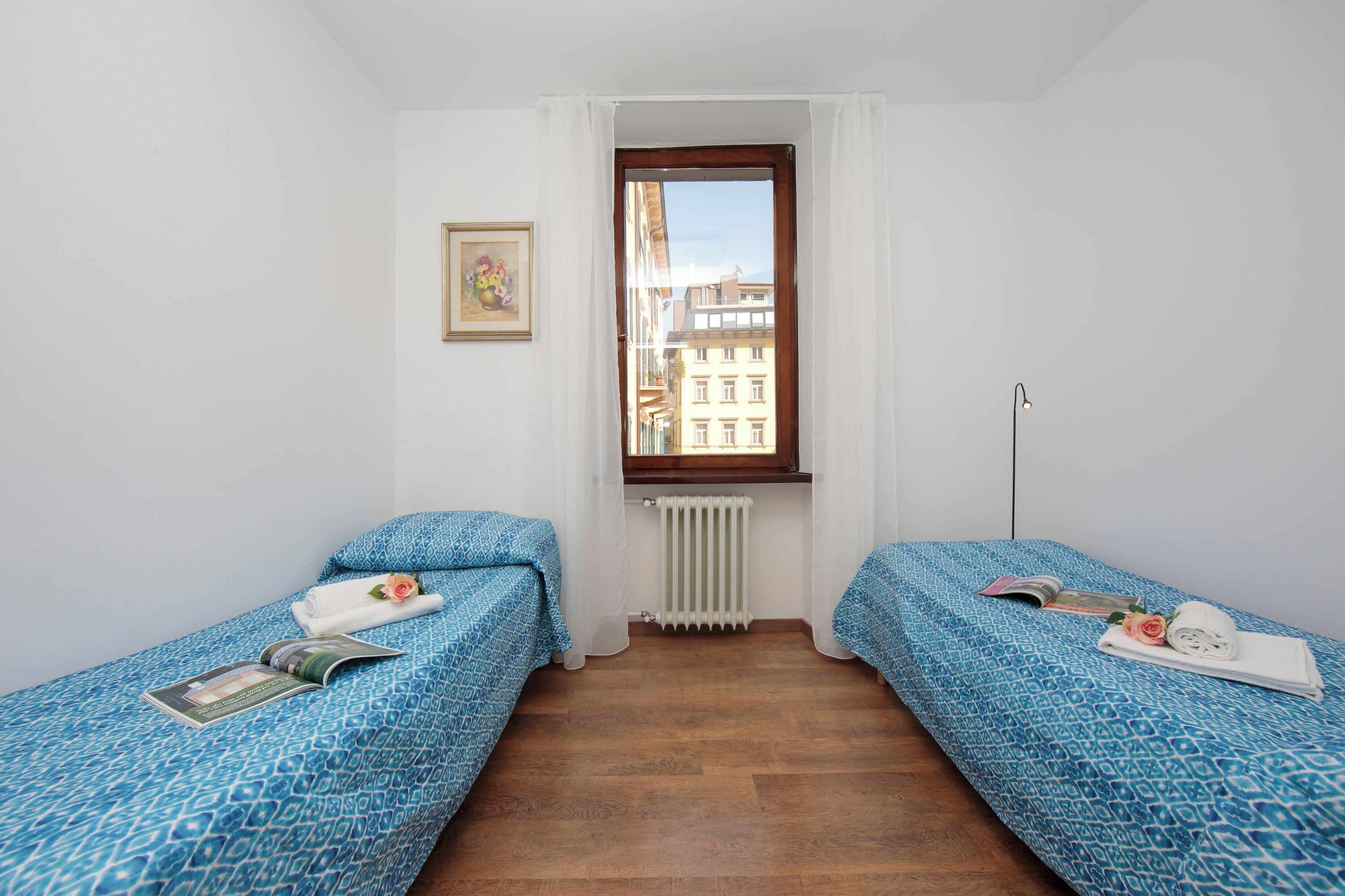 Ein Zimmer mit zwei Betten, einem Heizkörper, einem Fenster mit Vorhängen, einem Bild an der Wand und einer Stehlampe.