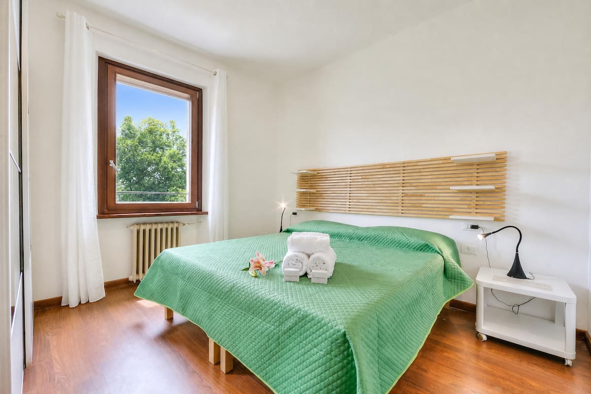 Une chambre avec un lit, une fenêtre avec des stores, un radiateur, une table de chevet avec une lampe et une petite plante.