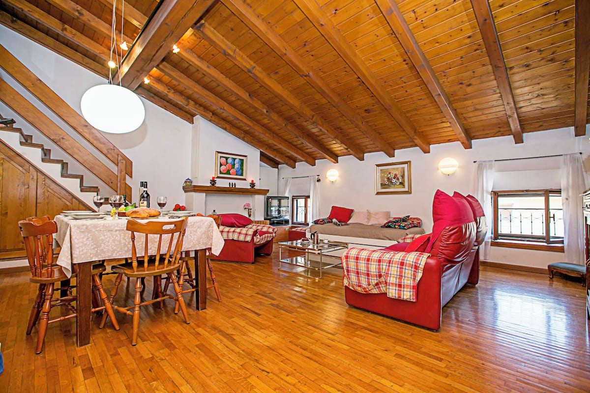 Un vaste salon doté de parquet et d’un haut plafond, comprenant un coin repas avec une table dressée pour quatre personnes, un canapé rouge et un téléviseur en arrière-plan.