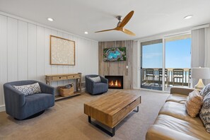 A living room with a fireplace, a ceiling fan, and a view of the beach through a sliding glass door.