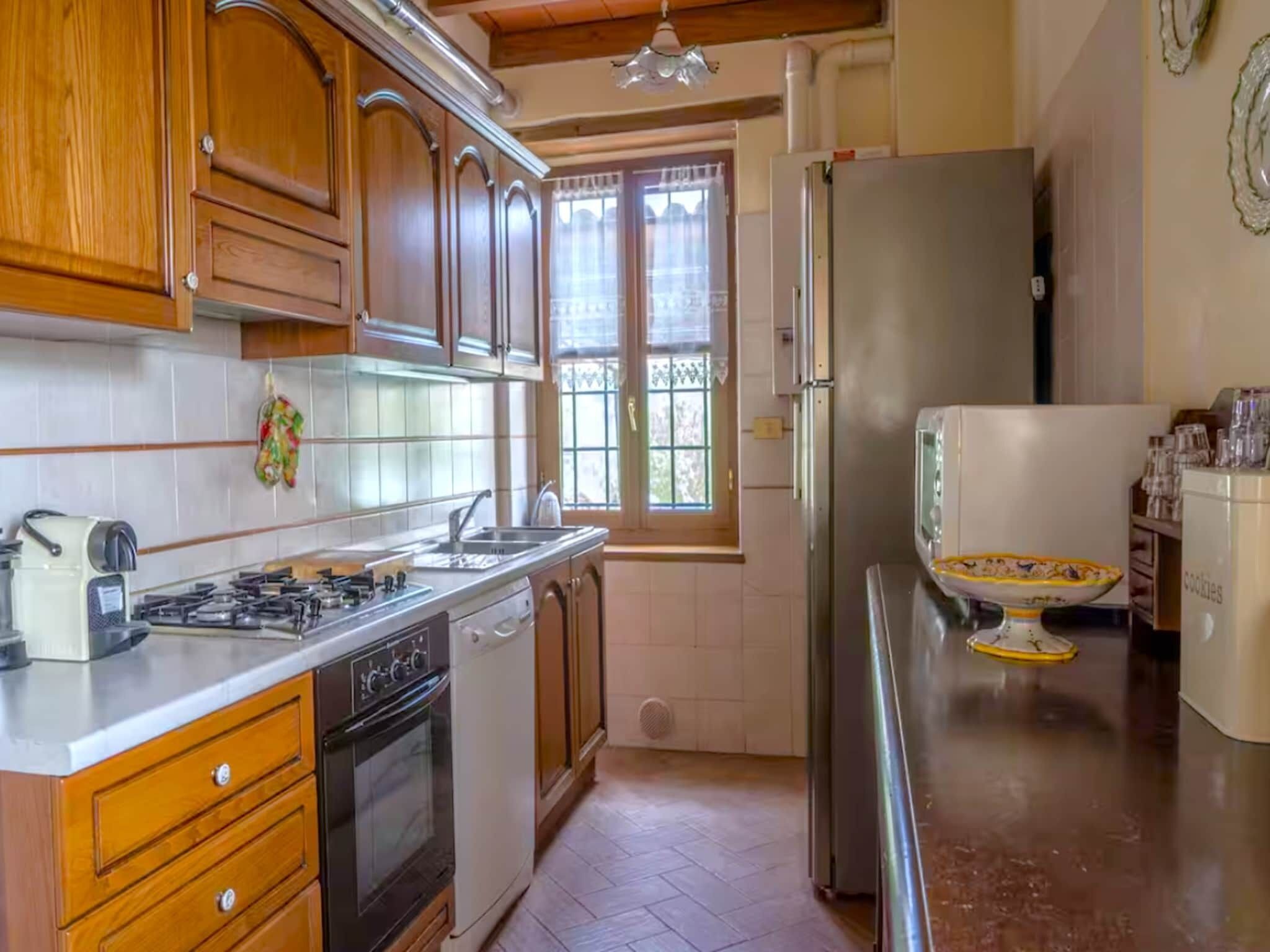 Une cuisine avec des meubles de rangement en bois, une cuisinière à gaz, un évier, un réfrigérateur et une fenêtre avec des rideaux.