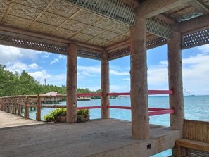 Um deck de madeira com telhado de palha, dando vista para a água.