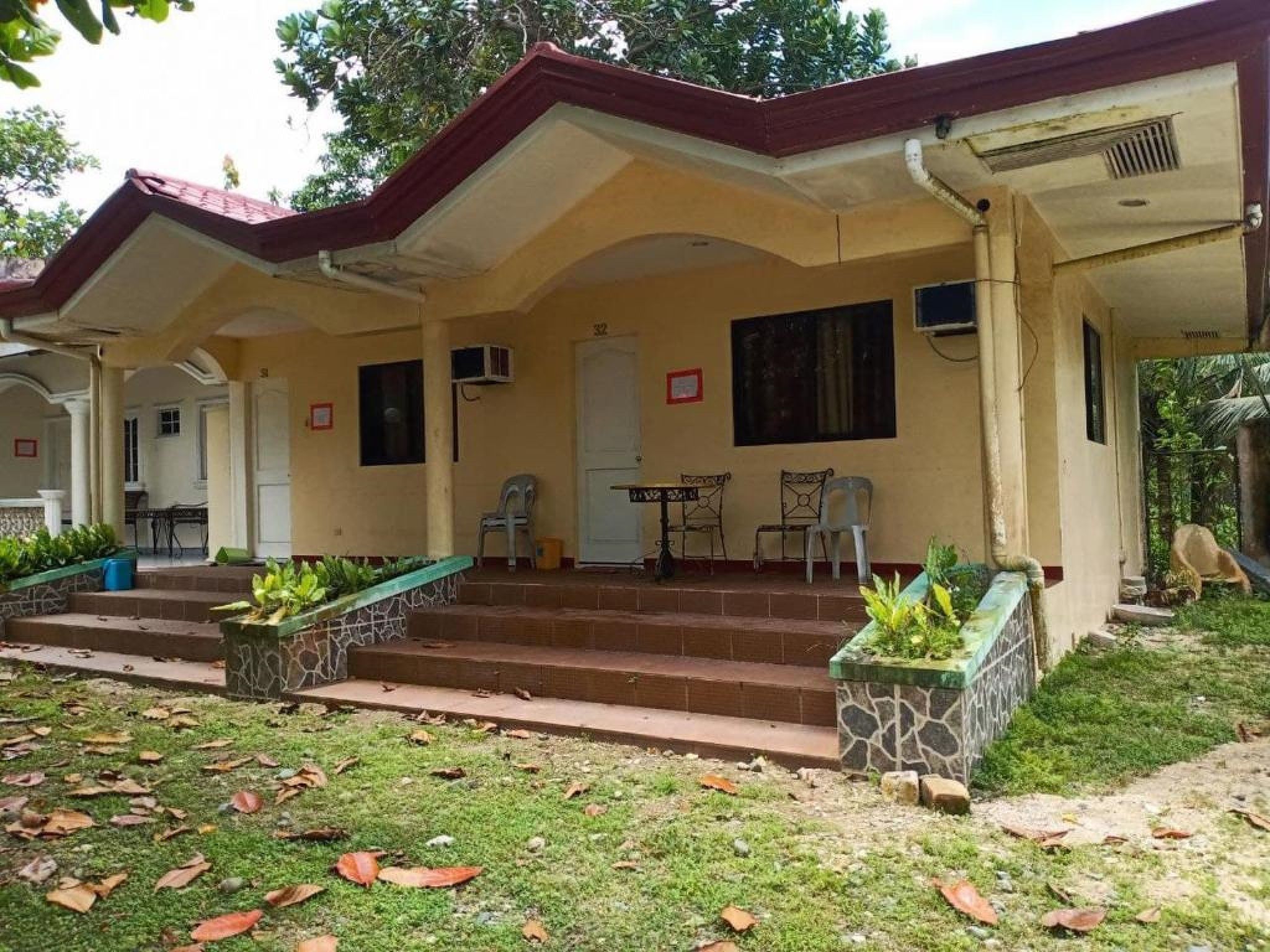 A two-story building with a covered porch, a dining area with a table and chairs, and a small garden area with steps leading to the entrance.