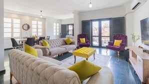 A spacious living room with a beige sofa, a purple armchair, and a yellow ottoman. There is a dining area with a table and chairs, and a large window with shutters.