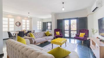 A spacious living room with a beige sofa, a purple armchair, and a yellow ottoman. There is a dining area with a table and chairs, and a large window with shutters.