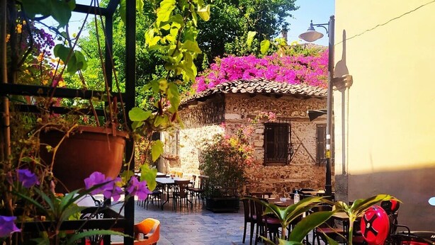 An outdoor patio area with tables and chairs, surrounded by potted plants and a stone building.