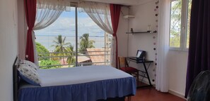 Ein Schlafzimmer mit Bett, Schreibtisch und Stuhl, ein großes Fenster mit Blick auf Palmen und Meer, sowie ein Regal mit Dekoration.