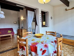 A dining area with a round wooden table, chairs, and a floral tablecloth. There is a sofa and a washing machine in the background.