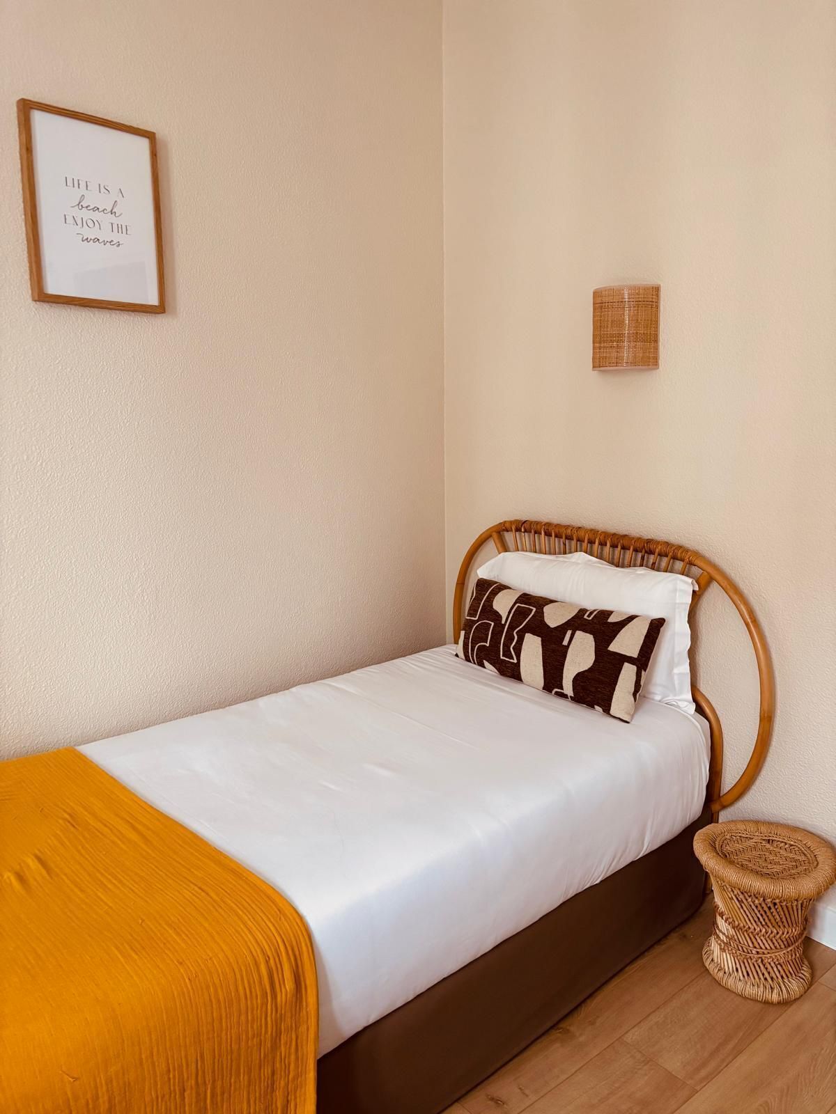 Een enkel bed met houten frame, witte beddengoed en een mosterdgele deken. Er staat een klein geweven kruk naast het bed en een ingelijst citaat hangt aan de muur.
