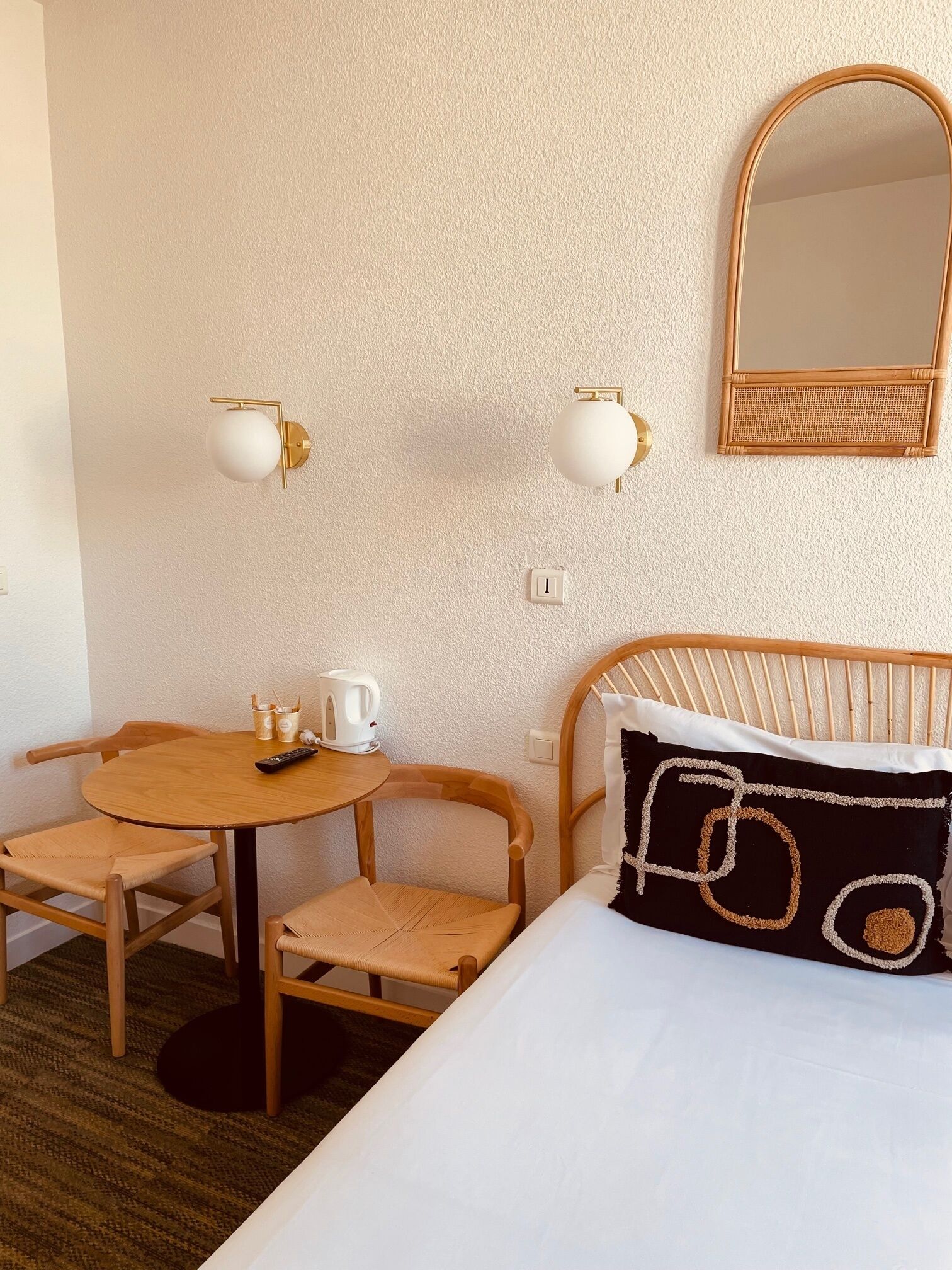 Une chambre d’hôtel avec une table ronde en bois, deux chaises, un lit avec un oreiller à motifs noir et blanc, un luminaire rond fixé au mur et un miroir sur le mur.