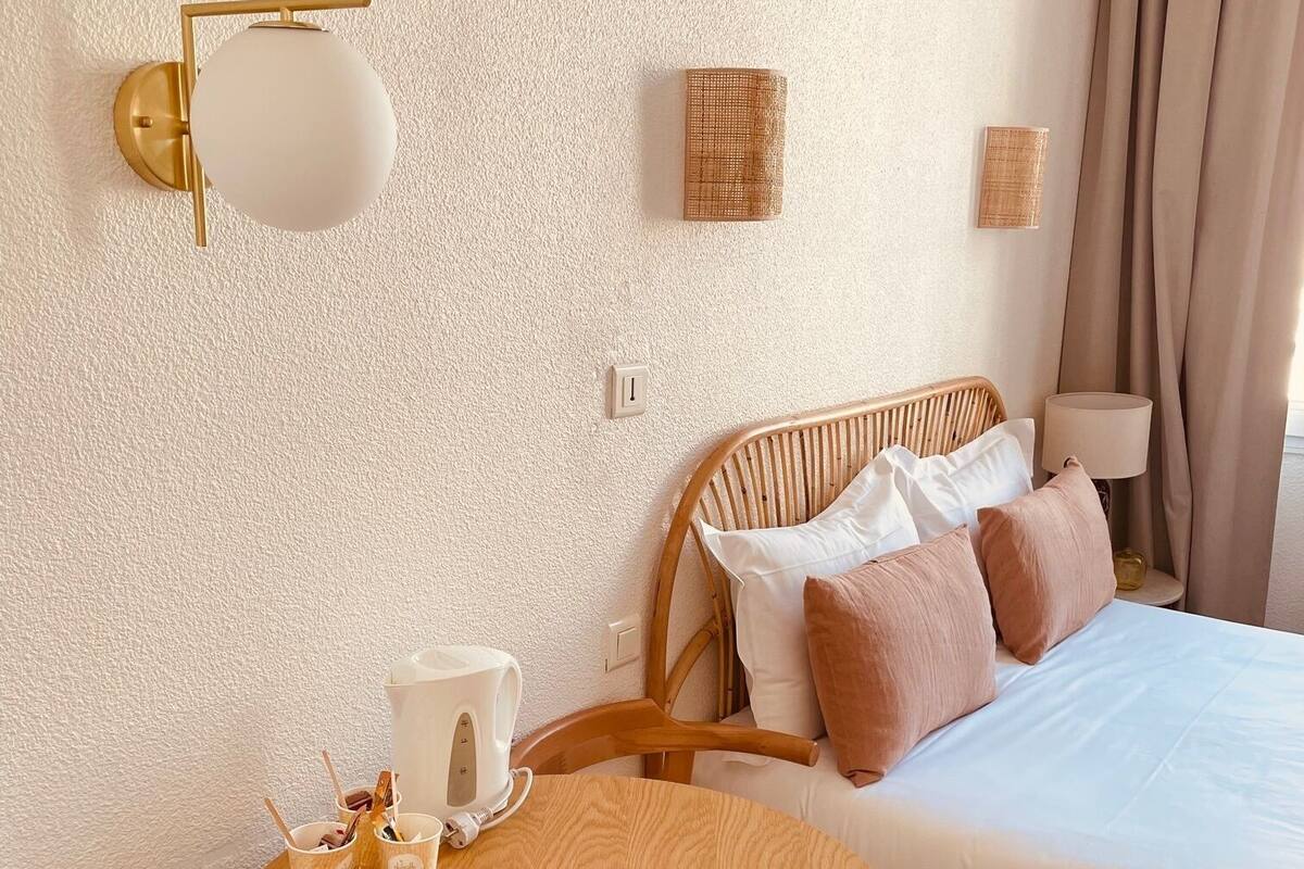 A hotel room with a bed, a round wooden table with cups and a kettle, a wall-mounted lamp, and a wicker chair.