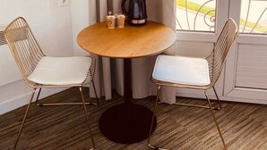 A room with a round wooden table, two gold wire chairs, a black coffee pot, and a window with a view of a building and trees. - Hôtel Le Britannic (Saint-Malo)