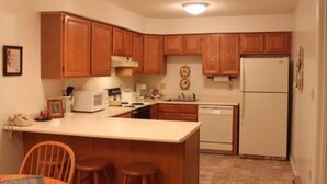 A compact kitchen with wooden cabinets, a white refrigerator, a microwave, and a sink. There are two stools at the counter and a small table with chairs. - Great vacation condo w/views of the mountains  #214 (Lincoln)